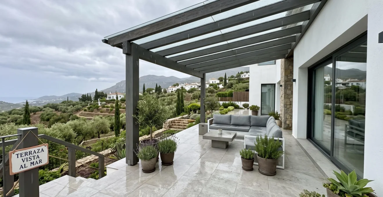Terraza contemporánea española con pérgola de doble inclinación instalada bajo cielo nublado con lluvia ligera visible, vegetación mediterránea al fondo