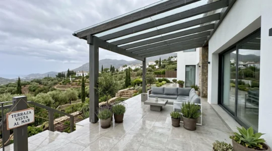 Terraza contemporánea española con pérgola de doble inclinación instalada bajo cielo nublado con lluvia ligera visible, vegetación mediterránea al fondo